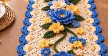 A crochet table runner with blue, yellow, and white 3D flowers and green leaves on a cream lacy base, finished with a blue scalloped edge.