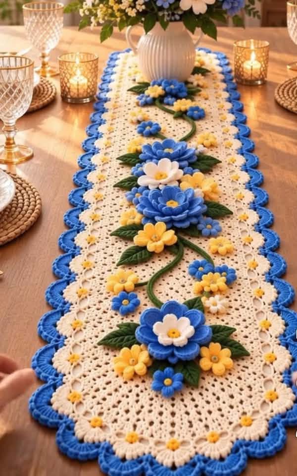 A crochet table runner with blue, yellow, and white 3D flowers and green leaves on a cream lacy base, finished with a blue scalloped edge.