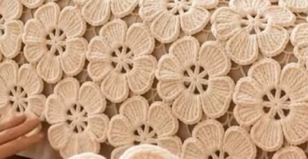 A handmade crochet bedspread made up of cream-colored large flower motifs joined together, with a close-up of one motif held in hand.