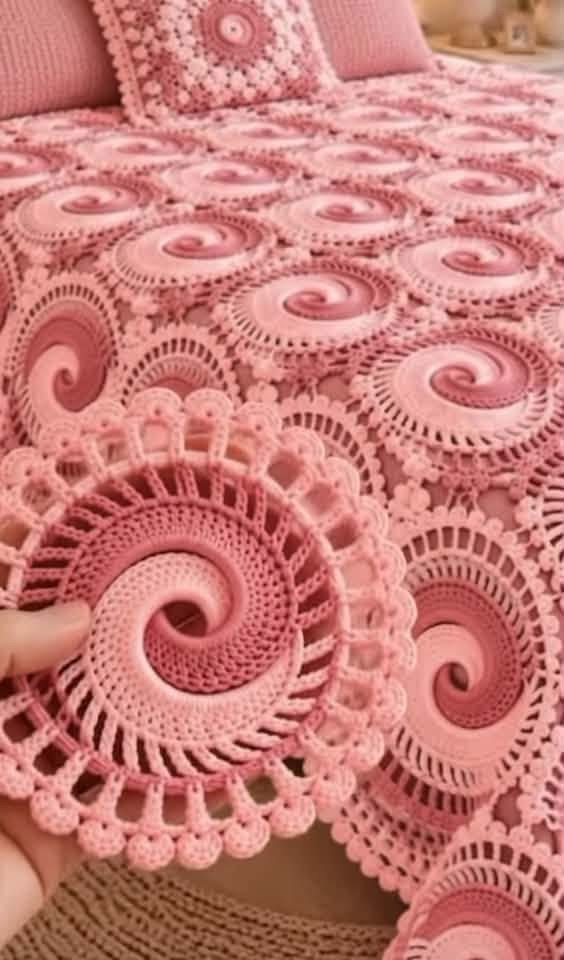 A pink crocheted blanket and pillow set featuring intricate swirling circular motifs, with a close-up of a single motif held in hand.