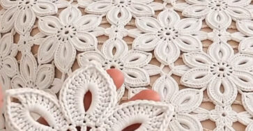 A hand holding a white crocheted flower motif in front of a bedspread made of similar joined motifs.
