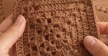 A hand holding a brown crochet granny square in front of a finished bedspread made from multiple granny squares.