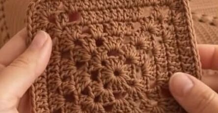 A hand holding a brown crochet granny square in front of a finished bedspread made from multiple granny squares.