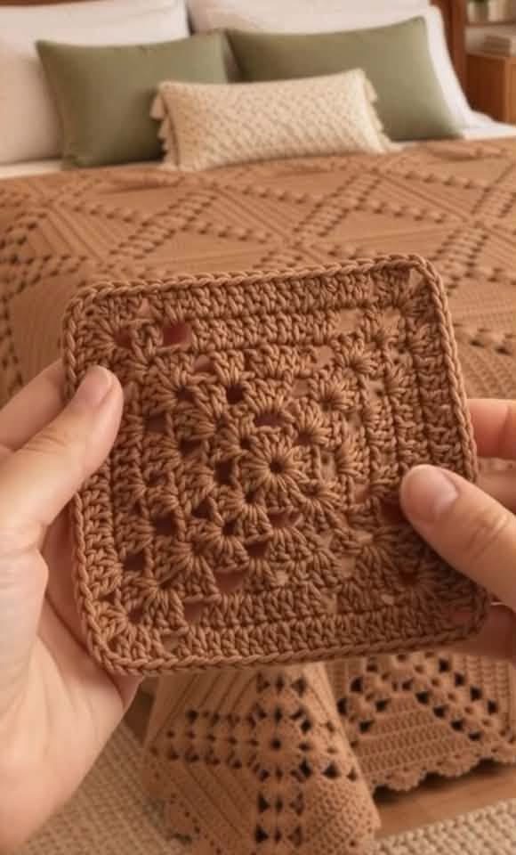 A hand holding a brown crochet granny square in front of a finished bedspread made from multiple granny squares.