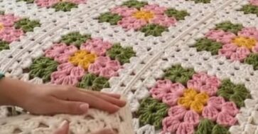 A bed with a crocheted blanket and pillow, both made of colorful floral granny squares in pink, yellow, green, and white tones.