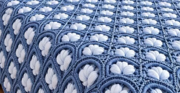 A plush crochet bedspread with intricate blue and white petal motifs joined as squares, displayed on a neatly made bed with matching pillows.