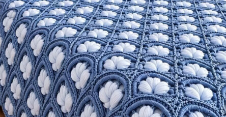 A plush crochet bedspread with intricate blue and white petal motifs joined as squares, displayed on a neatly made bed with matching pillows.