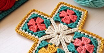 A crocheted cross made from vibrant granny squares with floral motifs, bordered in gold, surrounded by balls of yarn in various colors.