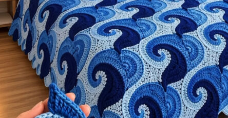 A bed covered with a crochet blanket made of blue layered spiral motifs, resembling ocean waves, with a person holding one motif in the foreground.