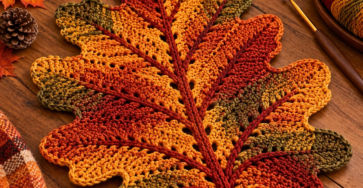 A large crocheted oak leaf placemat in fall colors of orange, red, yellow, and green with distinct veins, placed on a rustic wooden table.