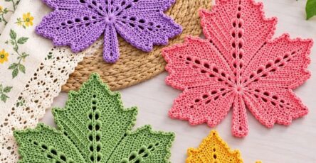 Four colorful crochet maple leaf coasters in purple, pink, green, and yellow displayed on a light table with flowers and decor.