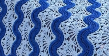 A crochet bedspread and matching pillows with blue and light blue wavy motifs and lacy fan designs, displayed on a bed.
