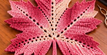 A crocheted maple leaf trivet in red and pink ombré shades, featuring pointed edges and textured ridges, displayed on a wooden table.
