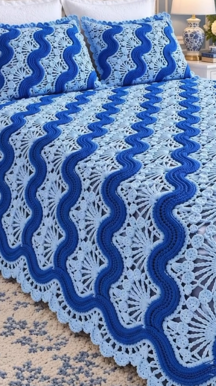 A crochet bedspread and matching pillows featuring alternating wavy stripes and intricate lacy fan motifs in light and royal blue yarn.