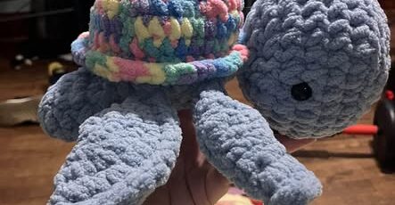 A hand holding a crocheted amigurumi turtle with a blue body and a chunky pastel-colored shell.
