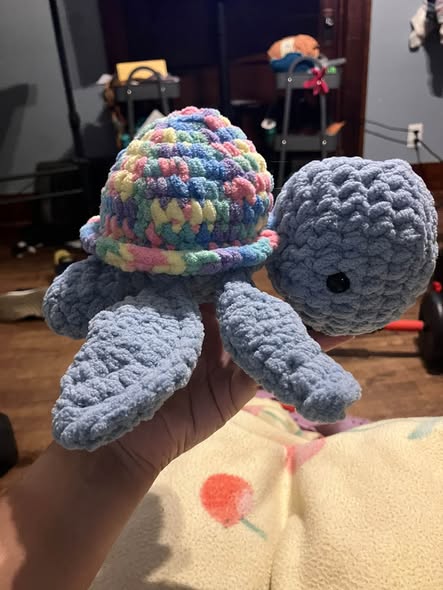 A hand holding a crocheted amigurumi turtle with a blue body and a chunky pastel-colored shell.
