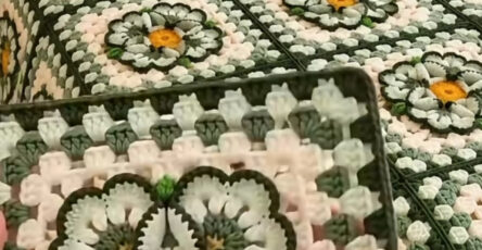 A hand holding a floral granny square motif in front of a matching crochet blanket and pillows with multicolor flower designs.
