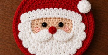 A round crochet appliqué showing a cute Santa Claus face with red hat and white beard on a wooden background.