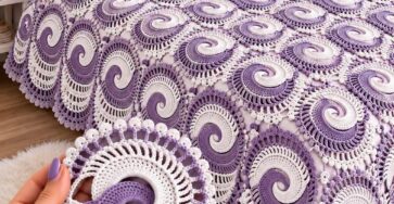 A crochet bedspread made of lavender and white spiral motifs with a decorative lacy border, shown on a bed in a cozy, elegant bedroom.
