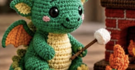 Crocheted green dragon plushie with wings, horns, and a marshmallow on a stick sitting by a fireplace.