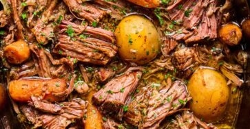 A slow cooker filled with tender pot roast, whole baby potatoes, carrots, and savory gravy, garnished with chopped parsley.