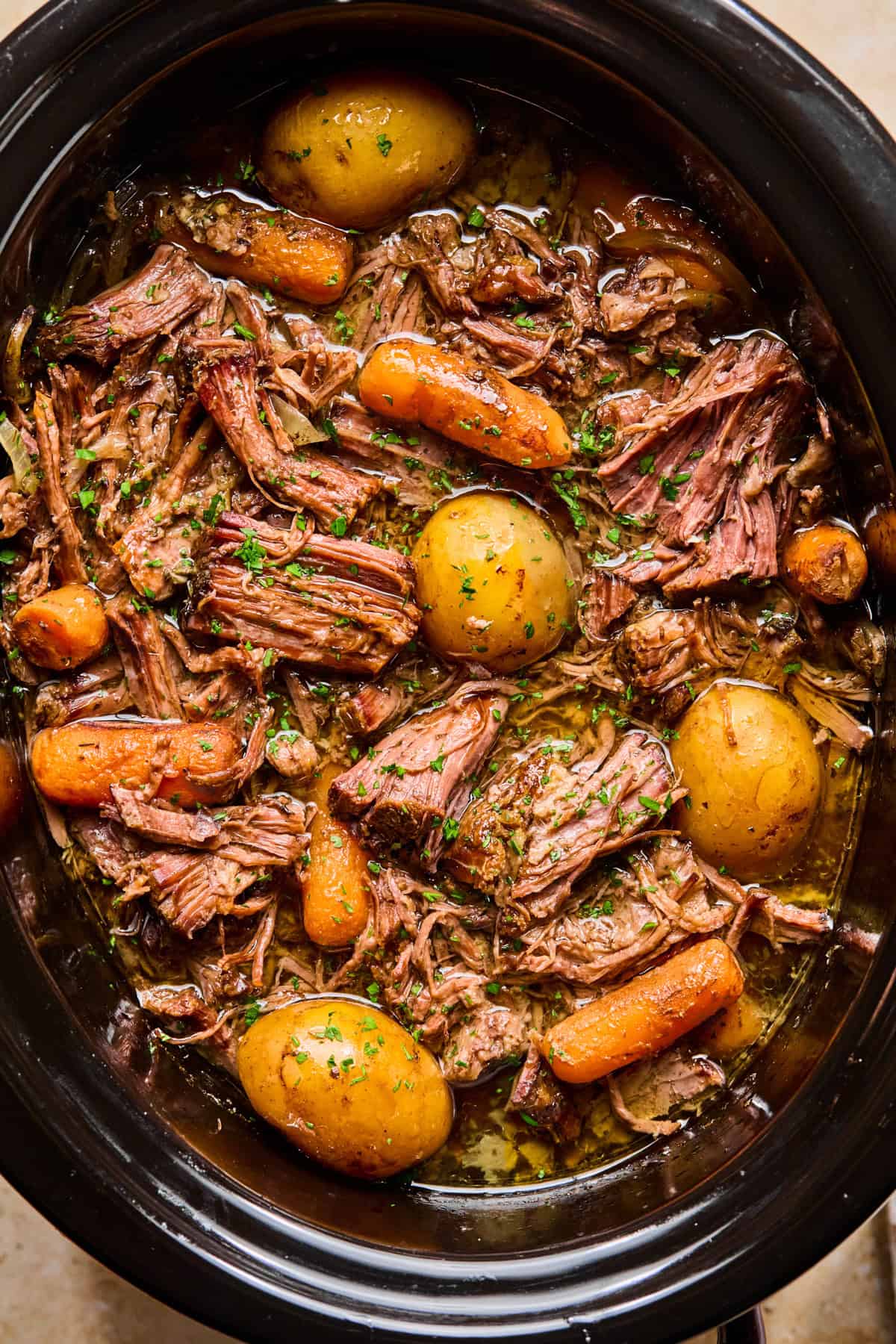 A slow cooker filled with tender pot roast, whole baby potatoes, carrots, and savory gravy, garnished with chopped parsley.