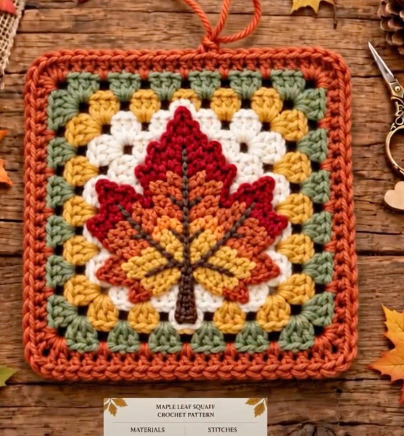 A square crochet piece featuring a vibrant maple leaf motif with fall colors, bordered in green, gold, and orange.