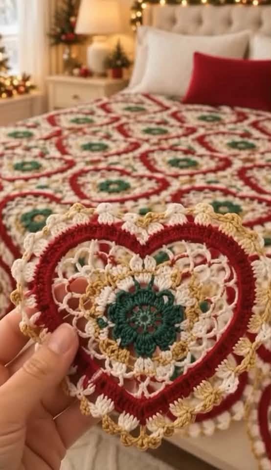 Hand holding a crochet heart motif in front of a matching heart motif crochet blanket on a bed, in red, green, white, and gold colors.