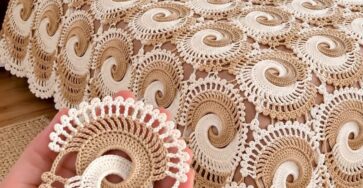 Hand holding a spiral crochet motif in cream and beige, with a bedspread made of joined spiral motifs in matching colors covering a bed.