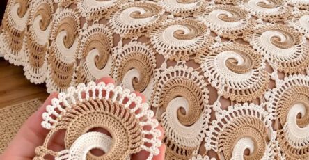 Hand holding a spiral crochet motif in cream and beige, with a bedspread made of joined spiral motifs in matching colors covering a bed.