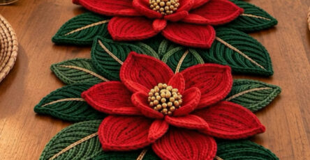 A festive crochet table runner featuring large red poinsettia flowers and layered green leaves arranged along a wooden dining table.