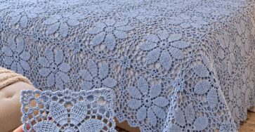 A light blue crochet bedspread with large floral motifs on a bed, with a close-up of one crocheted floral square held in a hand.