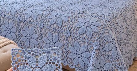 A light blue crochet bedspread with large floral motifs on a bed, with a close-up of one crocheted floral square held in a hand.