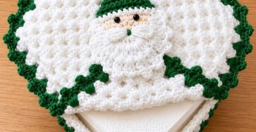 A crocheted Santa napkin holder featuring a white textured body with green trim and a Santa face motif.