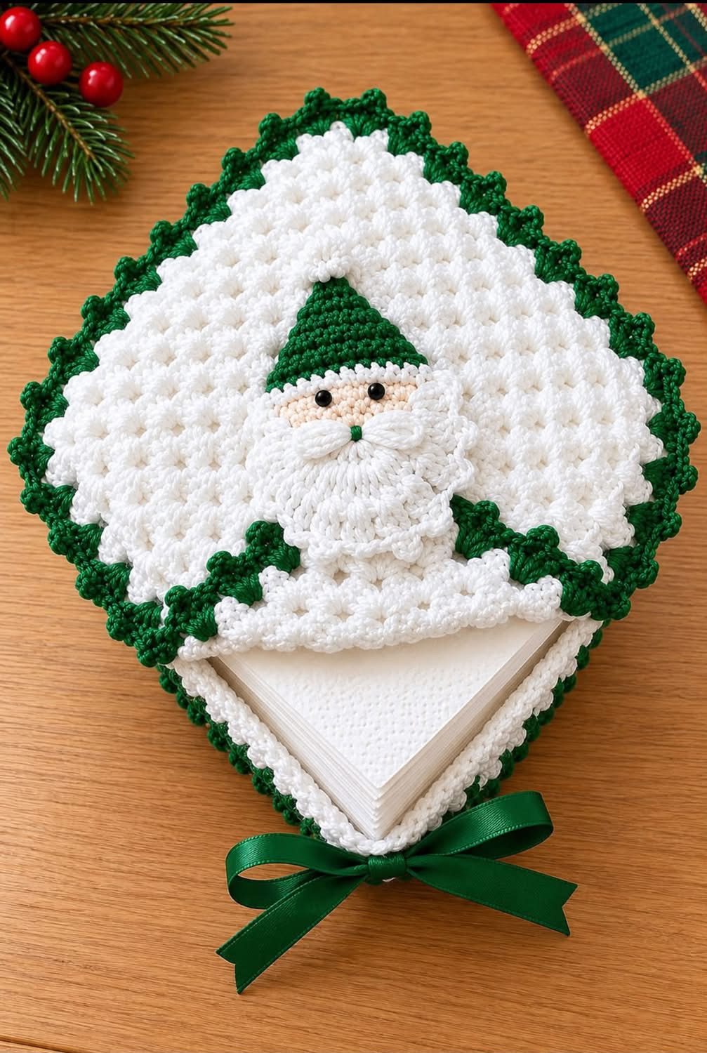 A crocheted Santa napkin holder featuring a white textured body with green trim and a Santa face motif.