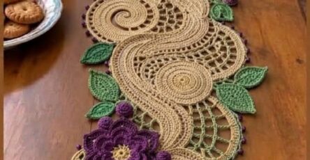 A long, decorative crochet table runner with swirling beige motifs, purple flowers, green leaves, and lace mesh accents arranged down the center of a table.