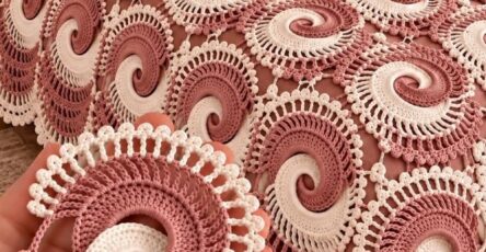 A hand holds a two-tone crochet swirl motif, shown alongside a bedspread made of many joined spiral motifs in dusty rose and ivory.