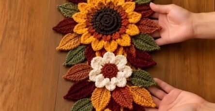 A crochet table runner with sunflowers, white flowers, and layered colored leaves in autumn hues.