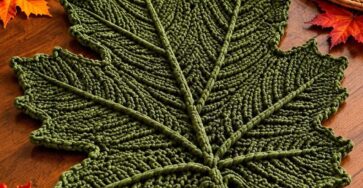 A crochet table mat shaped like a large green maple leaf, displayed on an autumn-themed dining table with pumpkins and fall leaves.