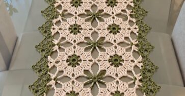 A crochet table runner with green and cream floral motifs arranged in lacy geometric patterns and bordered with a delicate green edge.