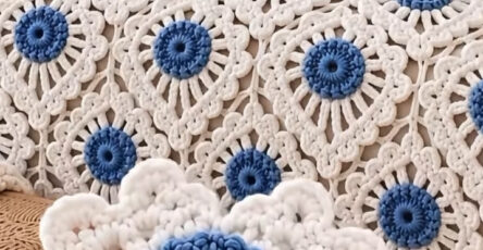 Hand holding a blue and white round crochet motif in front of a bedspread made from many identical motifs joined together.