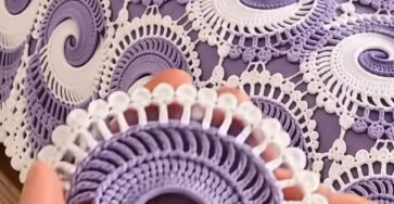 A close-up of a hand holding a lavender and white crochet spiral motif with an intricate lace edge, shown on a matching crochet bedspread.