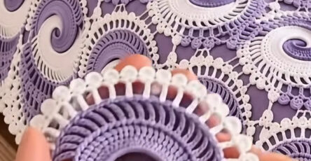 A close-up of a hand holding a lavender and white crochet spiral motif with an intricate lace edge, shown on a matching crochet bedspread.