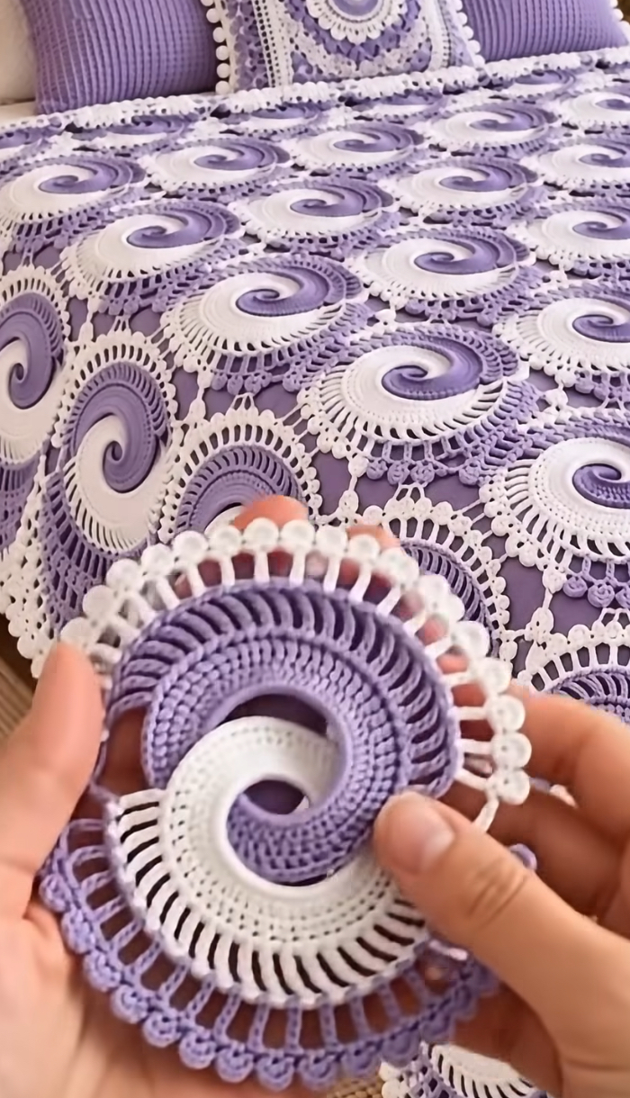 A close-up of a hand holding a lavender and white crochet spiral motif with an intricate lace edge, shown on a matching crochet bedspread.