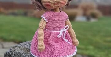 Amigurumi crochet doll with curly brown hair, wearing a pink dress, shoes, and a bow, seated outdoors.