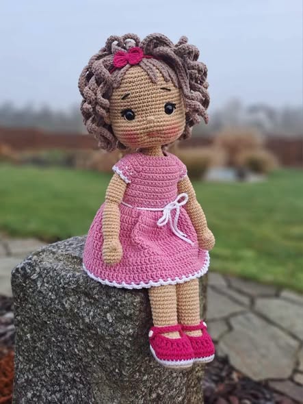 Amigurumi crochet doll with curly brown hair, wearing a pink dress, shoes, and a bow, seated outdoors.