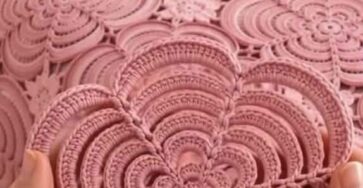 Hands holding a pink crochet flower motif in front of a bedspread made up of similar large motifs joined together.