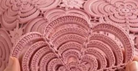 Hands holding a pink crochet flower motif in front of a bedspread made up of similar large motifs joined together.