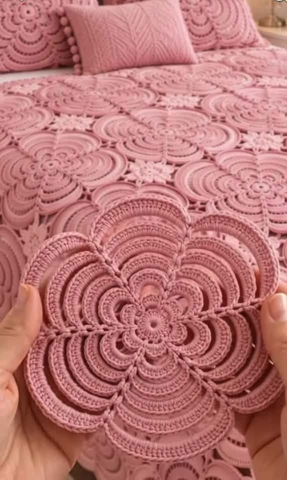 Hands holding a pink crochet flower motif in front of a bedspread made up of similar large motifs joined together.