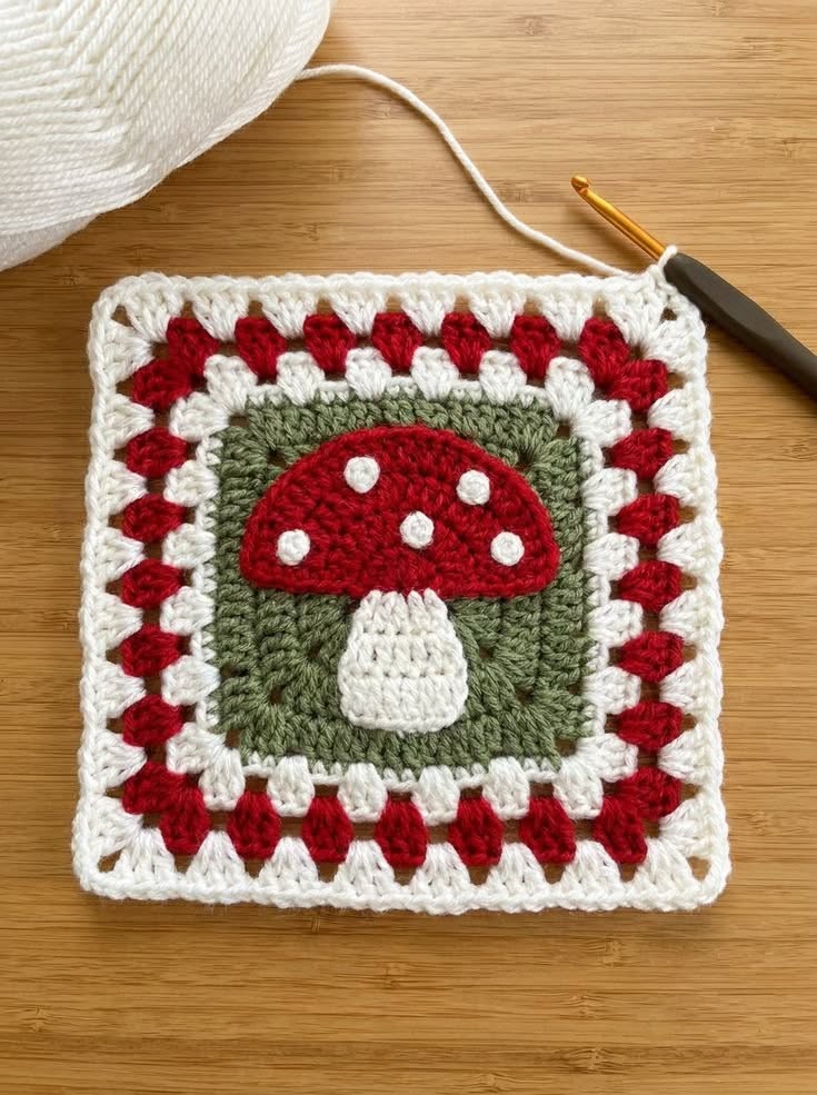 Crochet granny square with a red mushroom motif, white spots, green background, and alternating red and white granny borders.